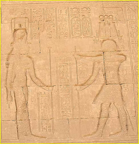A gauche, cette coiffe voquant la partie suprieure d'un sistre  naos, est bien videmment celle de la desse HATHOR, dont la prsence est rappele hiroglyphiquement parlant. Le bas-relief dmontre qu'elle fut jadis l'objet d'un martellement officiel, comme le visage du personnage de droite, dcid en haut lieu.